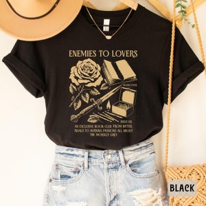 May include: Black t-shirt with a gold graphic design featuring a rose, a book, a dagger, and matches. The text on the shirt reads "Enemies to Lovers", "Villain Lover", "Issue 02", "An Exclusive Book Club From Bitter Rivals to Burning Passions All About The Morally Grey".
