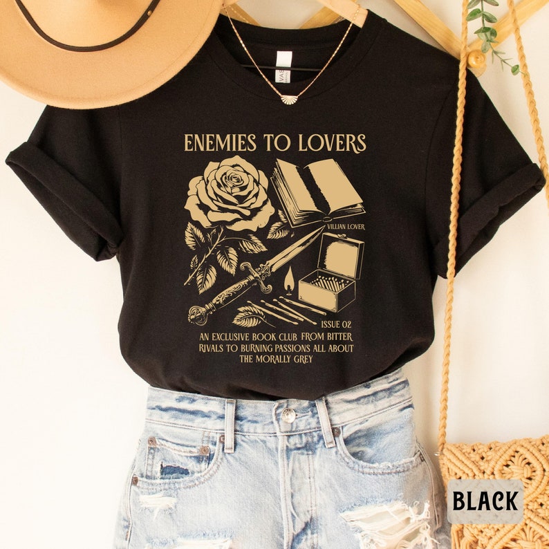 May include: Black t-shirt with a gold graphic design featuring a rose, a book, a dagger, and matches. The text on the shirt reads "Enemies to Lovers", "Villain Lover", "Issue 02", "An Exclusive Book Club From Bitter Rivals to Burning Passions All About The Morally Grey".