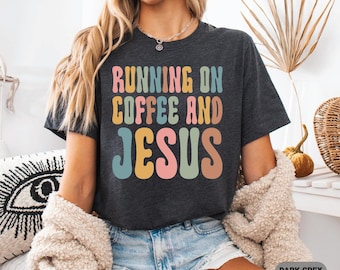 May include: Dark grey heather t-shirt with the text "RUNNING ON COFFEE AND JESUS" in colorful retro font. The shirt is a crew neck style and is being worn with denim shorts and a cream-colored cardigan.