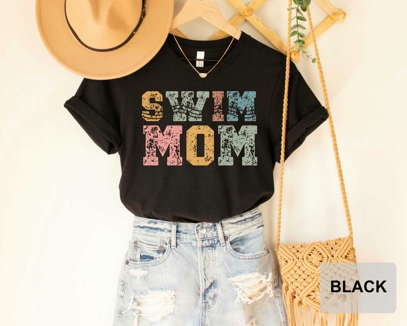 Pu&ograve; includere: Maglietta nera con la scritta "Swim Mom" in lettere colorate e consumate. La maglietta &egrave; esposta su un manichino con un cappello di paglia e una borsa intrecciata.