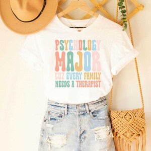 May include: White t-shirt with the text "Psychology Major Cuz Every Family Needs A Therapist" in colorful, retro-style lettering.