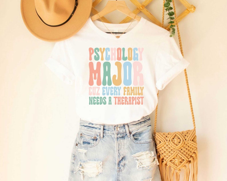 May include: White t-shirt with the text "Psychology Major Cuz Every Family Needs A Therapist" in colorful, retro-style lettering.