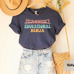 May include: Navy blue t-shirt with the text "Teacher? Educational Ninja" in colorful retro font. The shirt is styled with a straw hat, denim shorts, and a macrame bag.