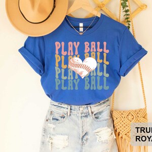 Peut inclure: Un t-shirt bleu avec le texte "Play Ball" dans un sch&eacute;ma de couleurs arc-en-ciel. Un ballon de baseball en forme de c&oelig;ur est au centre du texte. Le t-shirt est port&eacute; par une personne portant un short en jean.