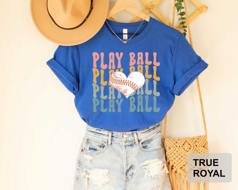 Peut inclure: Un t-shirt bleu avec le texte "Play Ball" dans un sch&eacute;ma de couleurs arc-en-ciel. Un ballon de baseball en forme de c&oelig;ur est au centre du texte. Le t-shirt est port&eacute; par une personne portant un short en jean.