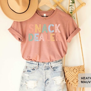 Puede incluir: Una camiseta de color malva con el texto "Snack Dealer" en letras de colores. La camiseta se muestra en una modelo que lleva pantalones cortos de mezclilla y un sombrero de paja.