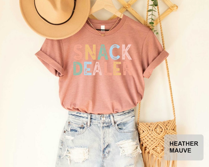 Puede incluir: Una camiseta de color malva con el texto "Snack Dealer" en letras de colores. La camiseta se muestra en una modelo que lleva pantalones cortos de mezclilla y un sombrero de paja.