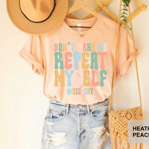 May include: A peach colored t-shirt with the text "Don't make me repeat myself #history" in colorful, retro-style lettering. The shirt is being modeled on a person wearing blue jeans and a brown hat.