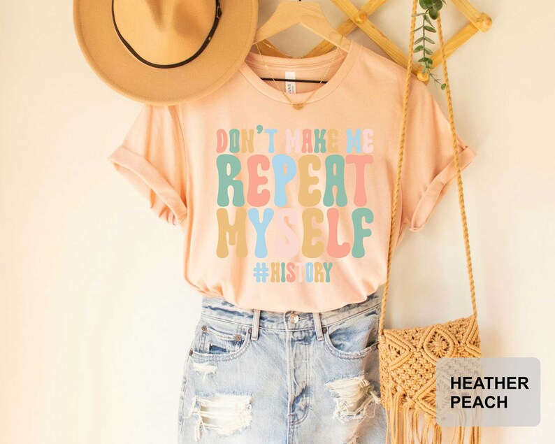 May include: A peach colored t-shirt with the text "Don't make me repeat myself #history" in colorful, retro-style lettering. The shirt is being modeled on a person wearing blue jeans and a brown hat.