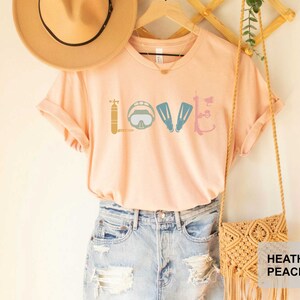 Peut inclure: Un t-shirt couleur p&ecirc;che avec le mot "LOVE" &eacute;crit en utilisant des graphismes d'&eacute;quipement de plong&eacute;e sous-marine. Le t-shirt est suspendu &agrave; un cintre en bois avec un sac en paille et un chapeau brun.