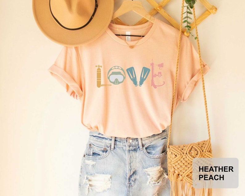 Peut inclure: Un t-shirt couleur p&ecirc;che avec le mot "LOVE" &eacute;crit en utilisant des graphismes d'&eacute;quipement de plong&eacute;e sous-marine. Le t-shirt est suspendu &agrave; un cintre en bois avec un sac en paille et un chapeau brun.
