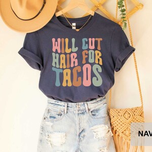 May include: A navy blue t-shirt with the text "Will Cut Hair For Tacos" in colorful, retro-style lettering. The shirt is shown on a model wearing denim shorts and a straw hat.