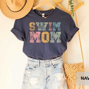 Pu&ograve; includere: Maglietta blu navy con una grafica in stile vintage che dice "Swim Mom" in lettere colorate.