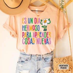 May include: A peach colored t-shirt with a colorful graphic of a flower with arms and legs. The text on the shirt reads "Es un dia hermoso para aprender cosas nuevas".