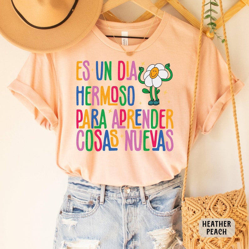 May include: A peach colored t-shirt with a colorful graphic of a flower with arms and legs. The text on the shirt reads "Es un dia hermoso para aprender cosas nuevas".
