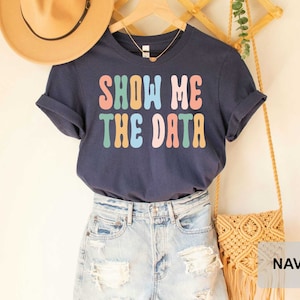May include: Navy blue t-shirt with the text "Show Me The Data" in rainbow colors. The shirt is displayed on a model wearing denim shorts and a straw bag.