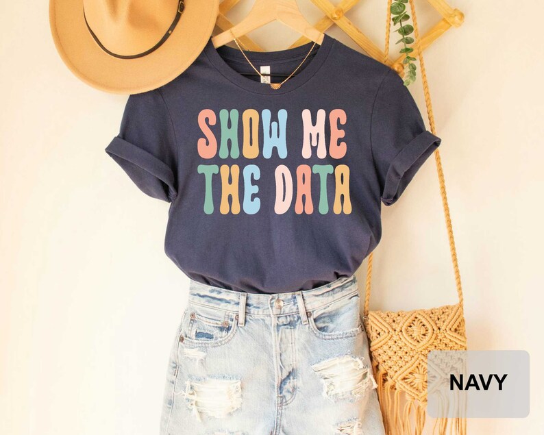 May include: Navy blue t-shirt with the text "Show Me The Data" in rainbow colors. The shirt is displayed on a model wearing denim shorts and a straw bag.