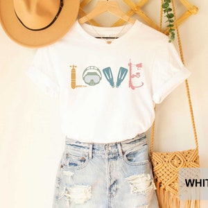 Peut inclure: T-shirt blanc avec un graphique d'&eacute;quipement de plong&eacute;e sous-marine qui &eacute;pelle le mot "LOVE". Le t-shirt est port&eacute; par une personne portant un short en jean et un chapeau de paille.