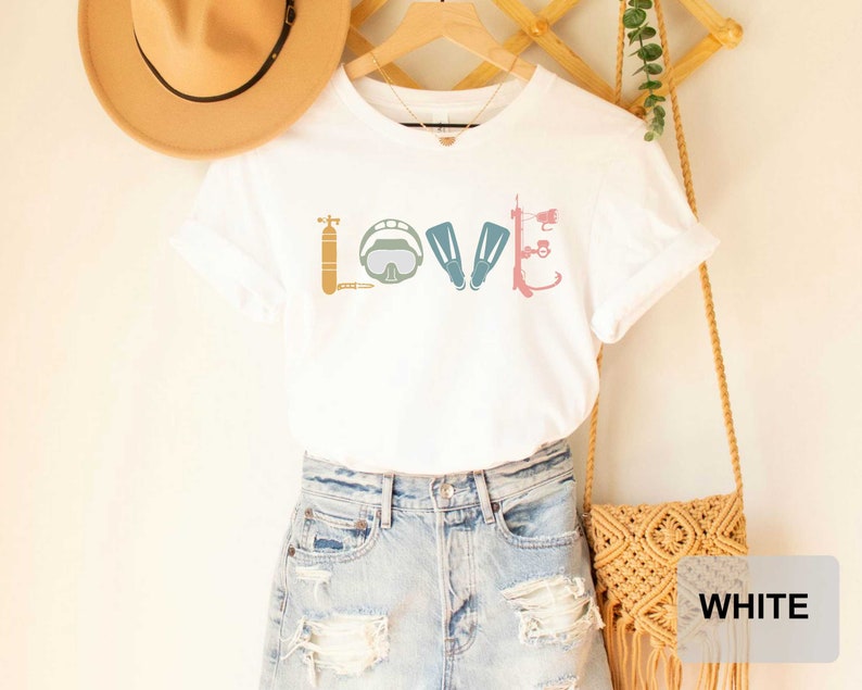 Peut inclure: T-shirt blanc avec un graphique d'&eacute;quipement de plong&eacute;e sous-marine qui &eacute;pelle le mot "LOVE". Le t-shirt est port&eacute; par une personne portant un short en jean et un chapeau de paille.