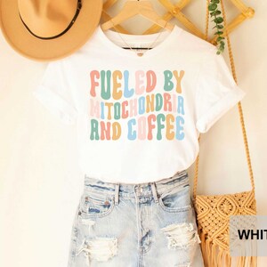 Op de afbeelding: Wit T-shirt met de tekst "Fueled by Mitochondria and Coffee" in kleurrijke, retro-stijl letters.