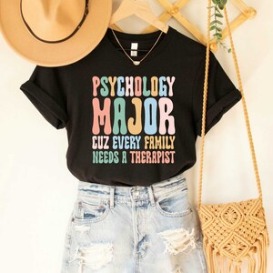 May include: Black t-shirt with the text "Psychology Major Cuz Every Family Needs A Therapist" in colorful retro font.