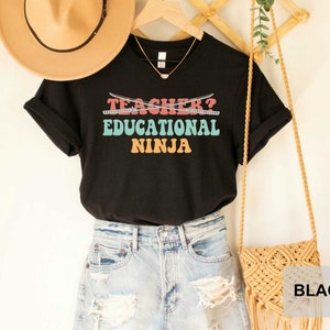 May include: Black t-shirt with the text "TEACHER? EDUCATIONAL NINJA" in colorful retro font with crossed swords above the text.