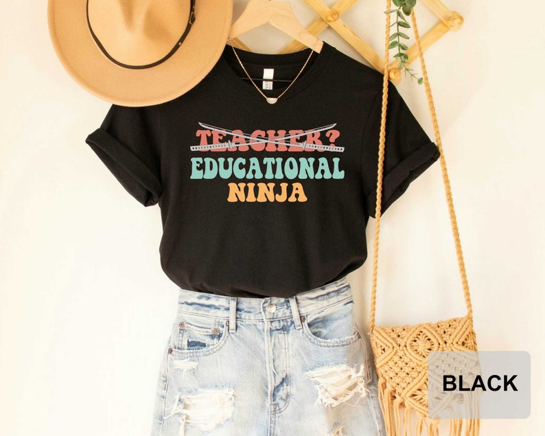 May include: Black t-shirt with the text "TEACHER? EDUCATIONAL NINJA" in colorful retro font with crossed swords above the text.