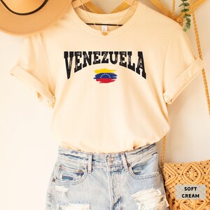 May include: A soft cream colored t-shirt with the word "VENEZUELA" in black distressed lettering and a stylized Venezuelan flag.