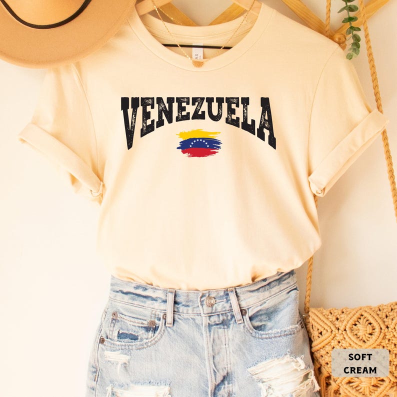 May include: A soft cream colored t-shirt with the word "VENEZUELA" in black distressed lettering and a stylized Venezuelan flag.