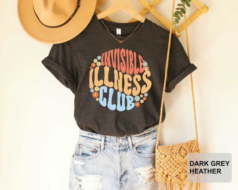 K&ouml;nnte beinhalten: Dunkelgraues T-Shirt mit einem bunten Aufdruck, auf dem "Invisible Illness Club" steht, umgeben von Blumen.