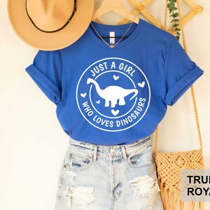 Puede incluir: Una camiseta azul real con un gr&aacute;fico blanco de un dinosaurio y el texto "Just a girl who loves dinosaurs".