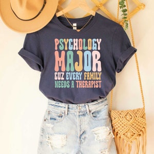 May include: A navy blue t-shirt with the text "Psychology Major Cuz Every Family Needs A Therapist" in colorful, retro-style lettering.