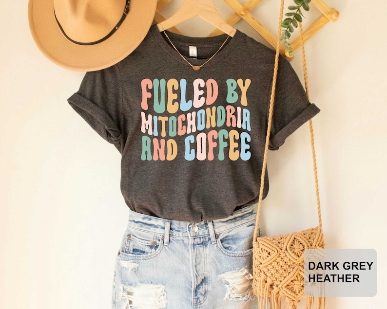 Op de afbeelding: Donkergrijs heather t-shirt met de tekst "Fueled by Mitochondria and Coffee" in regenboogkleuren.