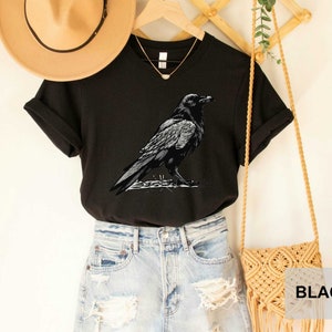 May include: Black t-shirt with a graphic of a crow on a branch. The shirt is shown on a model wearing denim shorts and a straw hat.