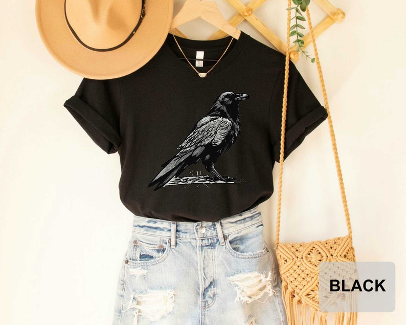 May include: Black t-shirt with a graphic of a crow on a branch. The shirt is shown on a model wearing denim shorts and a straw hat.