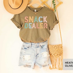 Puede incluir: Una camiseta verde con el texto "Snack Dealer" en letras de colores. La camiseta se combina con pantalones cortos de mezclilla y un bolso de paja con flecos.