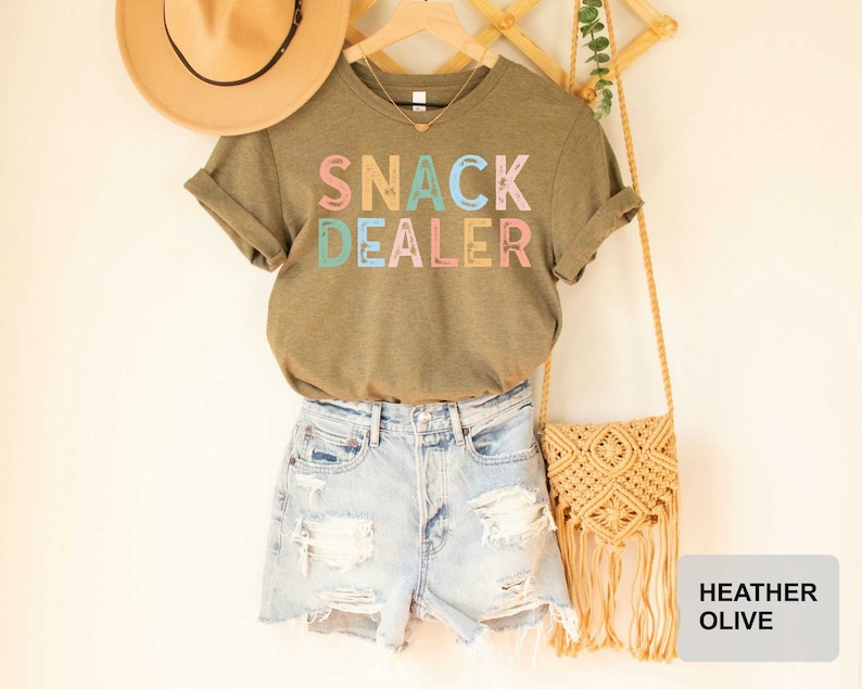Puede incluir: Una camiseta verde con el texto "Snack Dealer" en letras de colores. La camiseta se combina con pantalones cortos de mezclilla y un bolso de paja con flecos.