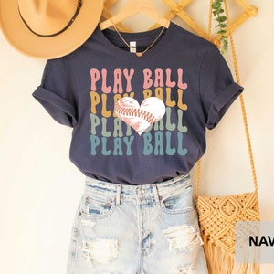 Peut inclure: T-shirt bleu marine avec un graphique de baseball en forme de c&oelig;ur et le texte "PLAY BALL" r&eacute;p&eacute;t&eacute; dans un sch&eacute;ma de couleurs arc-en-ciel.