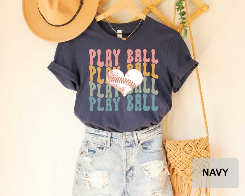 Peut inclure: T-shirt bleu marine avec un graphique de baseball en forme de c&oelig;ur et le texte "PLAY BALL" r&eacute;p&eacute;t&eacute; dans un sch&eacute;ma de couleurs arc-en-ciel.