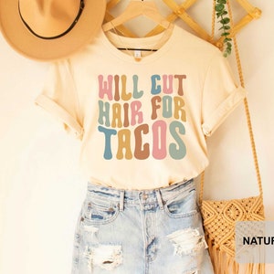 May include: A light beige t-shirt with the text "WILL CUT HAIR FOR TACOS" in colorful, retro-style lettering. The shirt is displayed on a model wearing denim shorts and a straw hat.