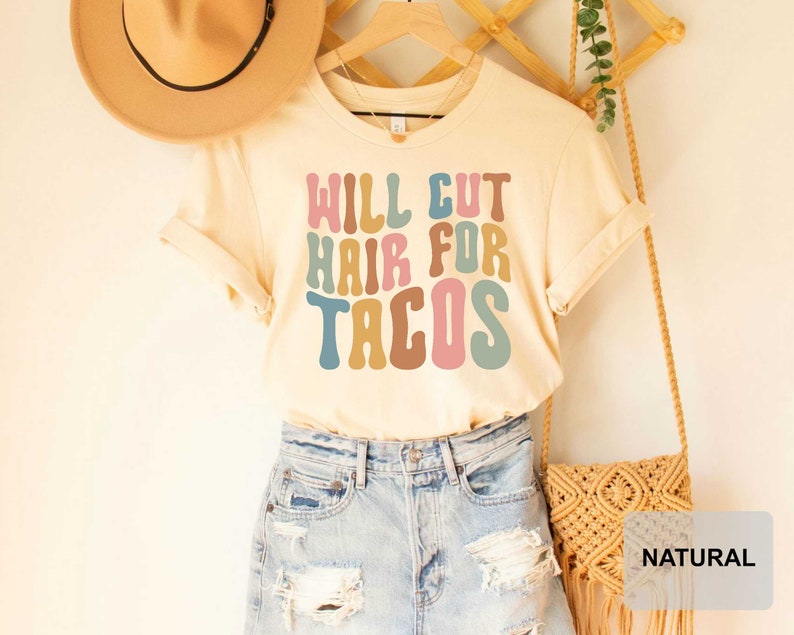 May include: A light beige t-shirt with the text "WILL CUT HAIR FOR TACOS" in colorful, retro-style lettering. The shirt is displayed on a model wearing denim shorts and a straw hat.