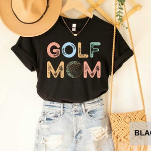 May include: Black t-shirt with the text "GOLF MOM" in colorful letters with a golf ball graphic. The shirt is being modeled with a straw hat and a woven bag.