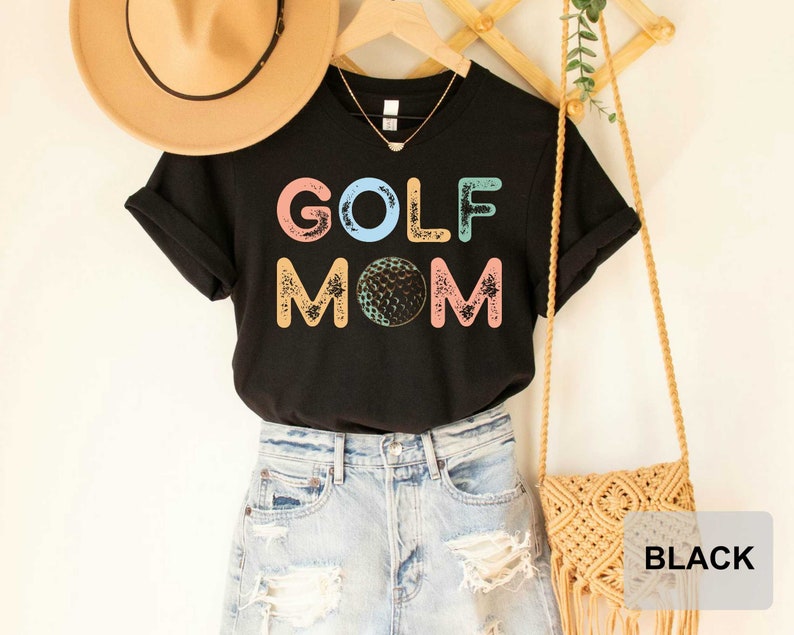 May include: Black t-shirt with the text "GOLF MOM" in colorful letters with a golf ball graphic. The shirt is being modeled with a straw hat and a woven bag.