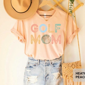 May include: A light pink t-shirt with the text "GOLF MOM" in colorful letters and a golf ball graphic. The shirt is shown on a model wearing blue jeans and a straw hat.