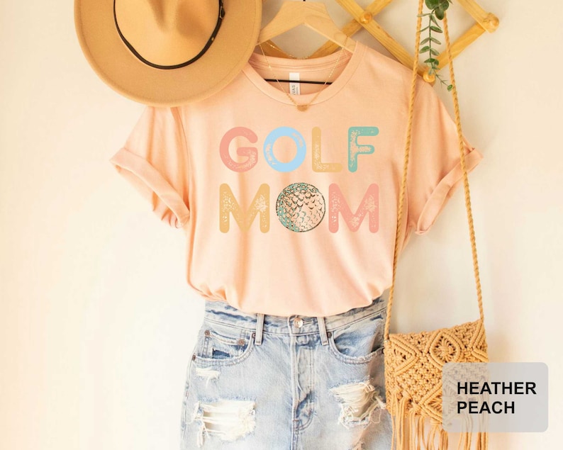 May include: A light pink t-shirt with the text "GOLF MOM" in colorful letters and a golf ball graphic. The shirt is shown on a model wearing blue jeans and a straw hat.