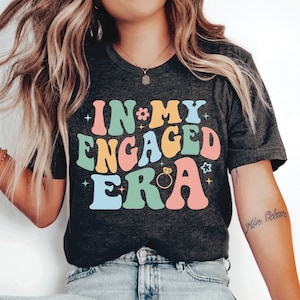 May include: A dark grey t-shirt with the text "IN MY ENGAGED ERA" in colorful retro lettering. The text includes a flower, a star, and a diamond ring.