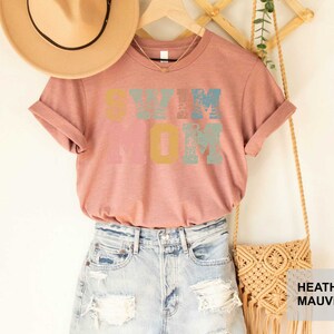 Pu&ograve; includere: Una maglietta color malva sfumato con il testo "Swim Mom" in lettere colorate e consumate. La maglietta &egrave; appesa a un supporto in legno con un cappello di paglia e una borsa in macram&eacute;.