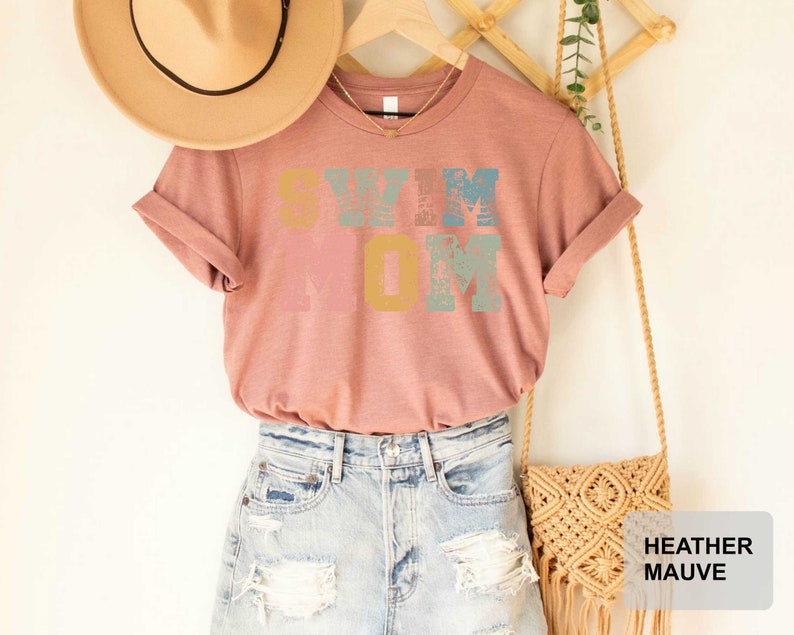 Pu&ograve; includere: Una maglietta color malva sfumato con il testo "Swim Mom" in lettere colorate e consumate. La maglietta &egrave; appesa a un supporto in legno con un cappello di paglia e una borsa in macram&eacute;.