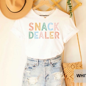 Puede incluir: Camiseta blanca con el texto "SNACK DEALER" en letras de colores. La camiseta est&aacute; en una percha y tiene un bolso de paja y un sombrero al lado.