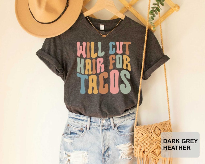 May include: A dark grey heather t-shirt with the text "Will Cut Hair For Tacos" in colorful retro font.